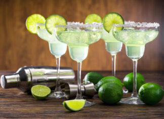 Margarita Prices Creeping Up Four glasses of freshly made Margarita cocktail decorated with limes on wooden table with shaker and limes.